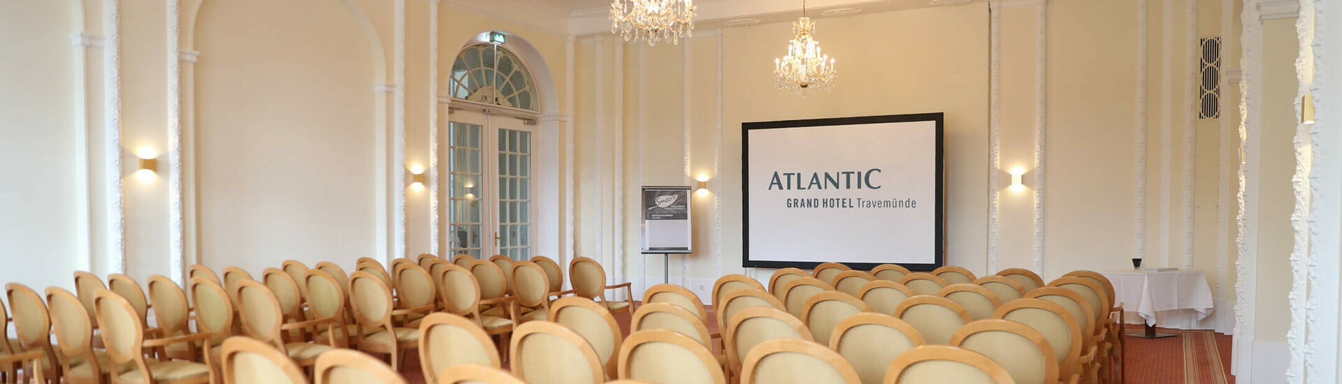 Tagungs- und Veranstaltungsraum "Bremen" Eleganter Konferenzraum mit Kronleuchtern, Reihen von Stühlen und einer Leinwand im ATLANTIC Grand Hotel Travemünde.