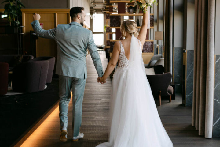 A wedding couple walk hand in hand through an elegant hotel corridor, the bride holding up a bouquet of flowers.