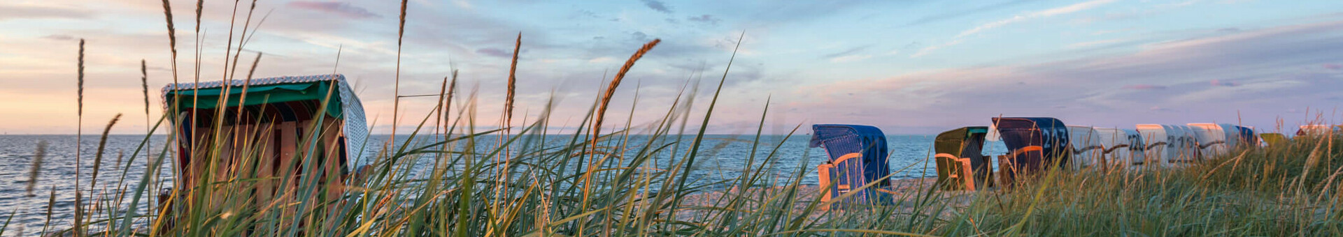 Strandkörbe am Meer bei Sonnenuntergang, umgeben von hohem Gras unter blauem Himmel.