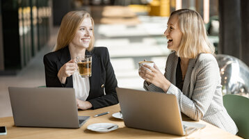 Two women are laughing and sitting in front of their laptops, holding cups of coffee.