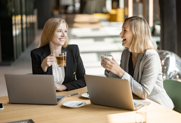 Zwei Frauen lachen und sitzen vor Laptops während sie Kaffee in der Hand halten.
