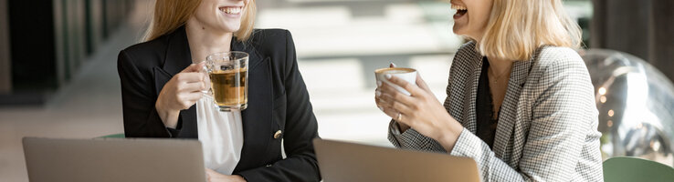 Two women are laughing and sitting in front of their laptops, holding cups of coffee.