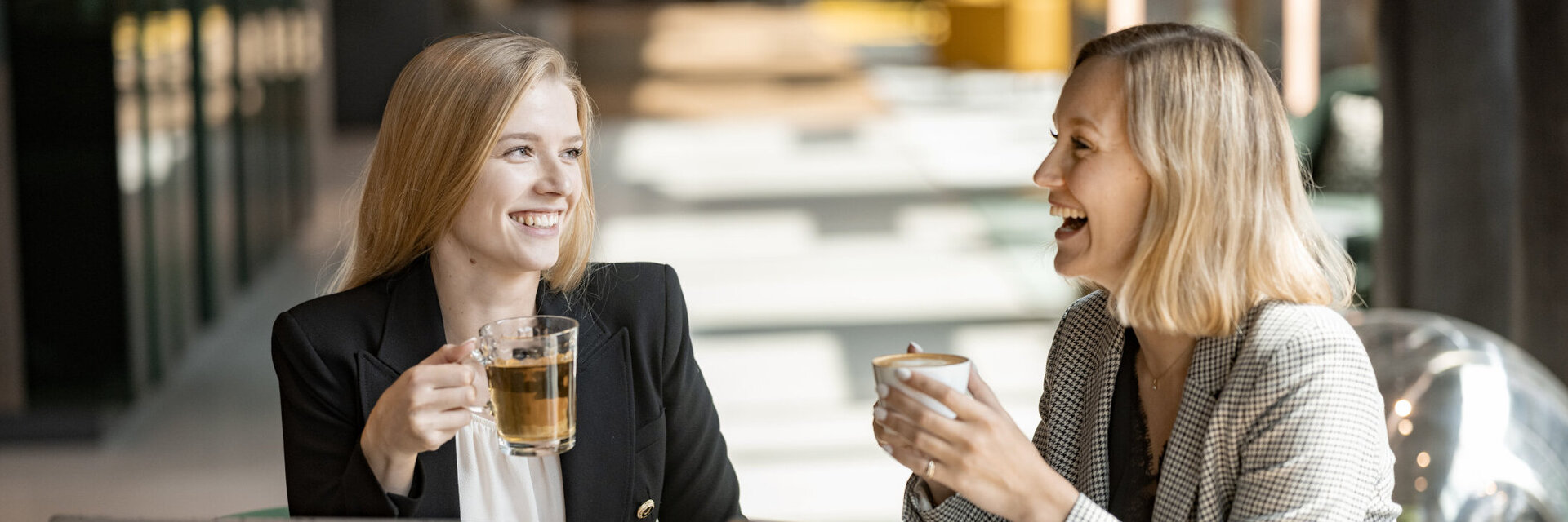 Two women are laughing and sitting in front of their laptops, holding cups of coffee.