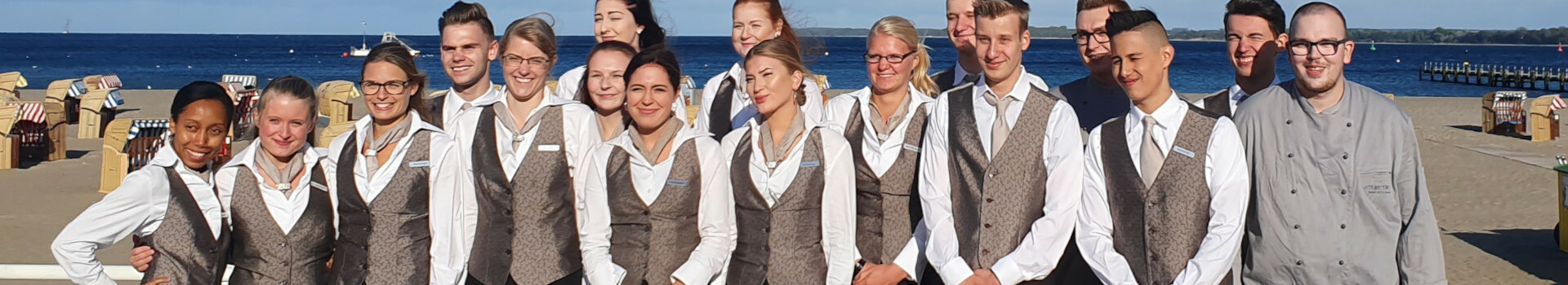 Hotel staff in uniforms smiling and posing on the beach in front of the blue sea and beach chairs.