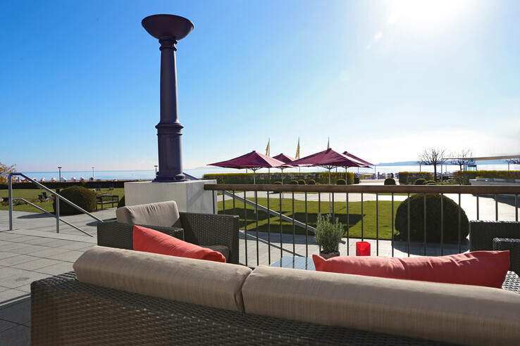 Terrasse mit gemütlichen Sitzgelegenheiten, Blick auf Rasen, rote Sonnenschirme und Meer im Hintergrund.