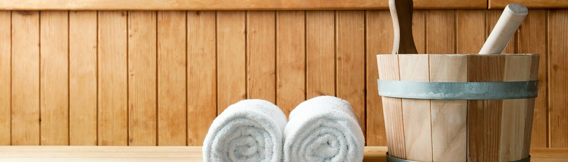 Wooden sauna with two rolled towels and a bucket with a ladle on a bench.