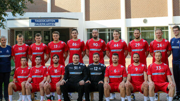 Eine Handballmannschaft in roten Trikots posiert vor einem Gebäude für ein Gruppenfoto.