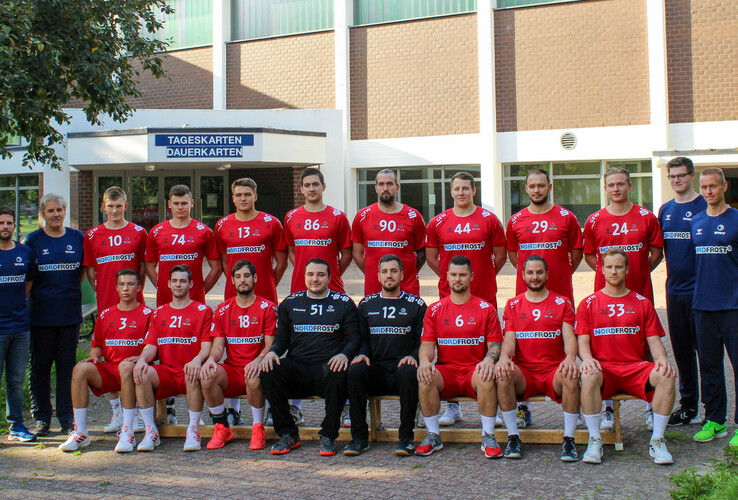 Eine Handballmannschaft in roten Trikots posiert vor einem Gebäude für ein Gruppenfoto.