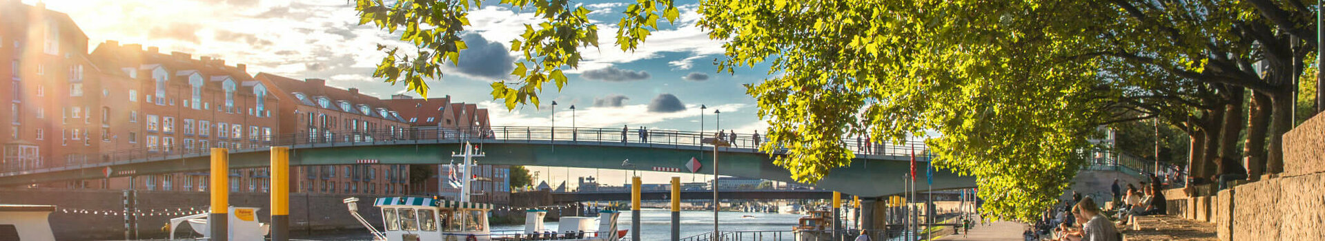 Sonniger Spazierweg an der Weser mit Blick auf Boote, Brücke und Bäume, nahe dem ATLANTIC Grand Hotel Bremen.