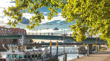 Sonniger Spazierweg an der Weser mit Blick auf Boote, Brücke und Bäume, nahe dem ATLANTIC Grand Hotel Bremen.