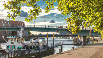 Sunny walk along the Weser with a view of boats, bridge and trees, close to the ATLANTIC Grand Hotel Bremen.