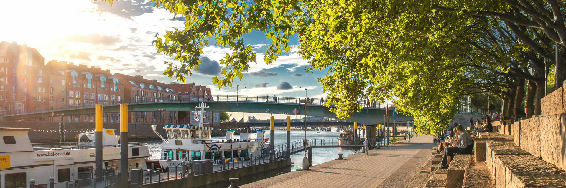 Sonniger Spazierweg an der Weser mit Blick auf Boote, Brücke und Bäume, nahe dem ATLANTIC Grand Hotel Bremen.