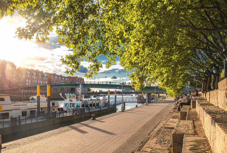 Sunny walk along the Weser with a view of boats, bridge and trees, close to the ATLANTIC Grand Hotel Bremen.