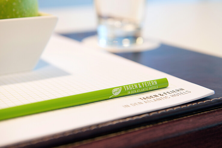 Grüner Stift auf Notizblock mit "Tagen & Feiern" Schriftzug, daneben Apfel in Schale und Wasserglas im Hintergrund.