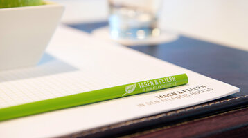 Grüner Stift auf Notizblock mit "Tagen & Feiern" Schriftzug, daneben Apfel in Schale und Wasserglas im Hintergrund.