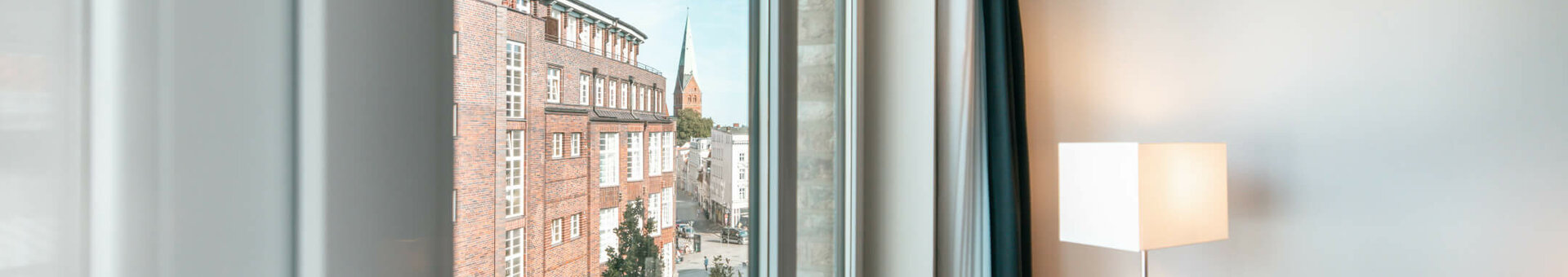 Blick aus einem Hotelzimmerfenster auf Lübecks Altstadt mit rotem Sessel und Stehlampe im Vordergrund.
