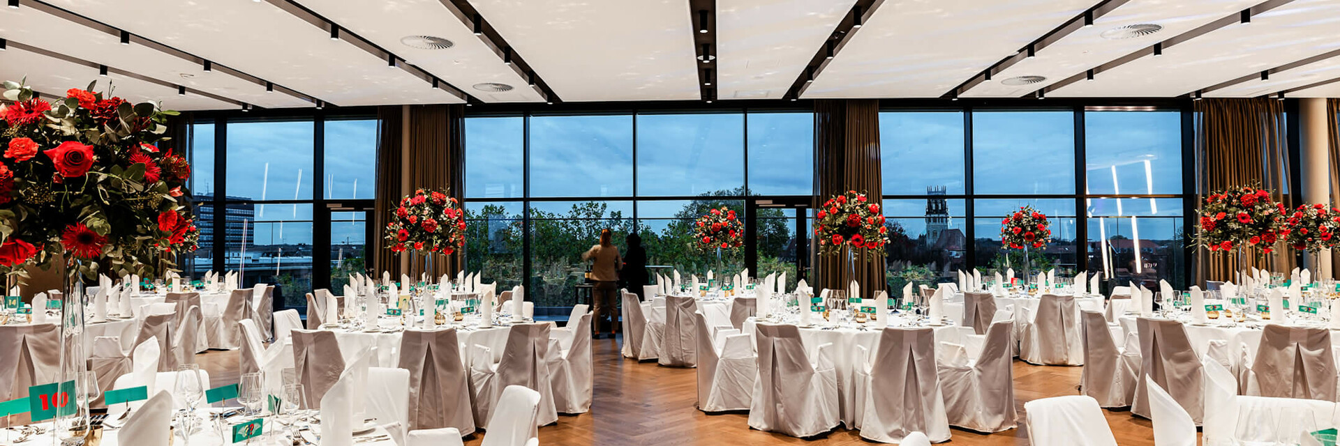Eleganter Bankettsaal im ATLANTIC Hotel Münster, mit weiß gedeckten Tischen, roten Blumenarrangements und Panoramafenstern.