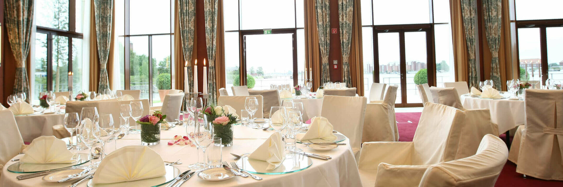 Elegant banquet hall in the ATLANTIC Hotel Wilhelmshaven, festively laid tables with a view of large windows and water.