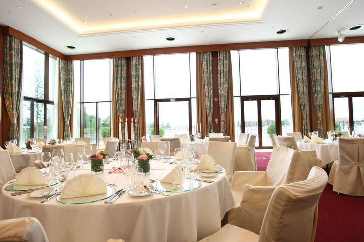 Elegant banquet hall in the ATLANTIC Hotel Wilhelmshaven, festively laid tables with a view of large windows and water.