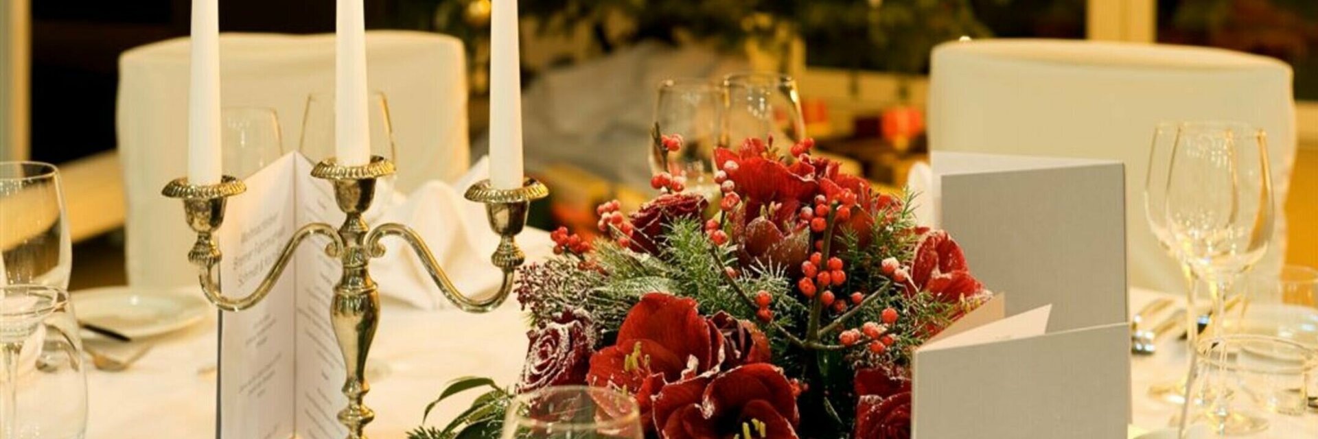 Festlich gedeckter Tisch mit Kerzenleuchter, roten Blumen und Weihnachtsbaum im Hintergrund.