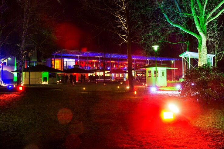 Nachtaufnahme des ATLANTIC Hotel Galopprennbahn, beleuchtet in bunten Farben mit Bäumen und Garten im Vordergrund.