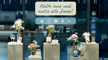 Fünf Aufsteller in Betonoptik, darauf jeweils ein kleiner Blumenstrauß und die Ausstellungsstücke, wie Kaffeebohnen, Duftstäbchen und eine Cocktailprobe.