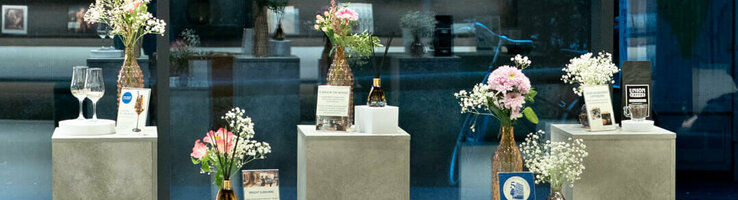 Fünf Aufsteller in Betonoptik, darauf jeweils ein kleiner Blumenstrauß und die Ausstellungsstücke, wie Kaffeebohnen, Duftstäbchen und eine Cocktailprobe.