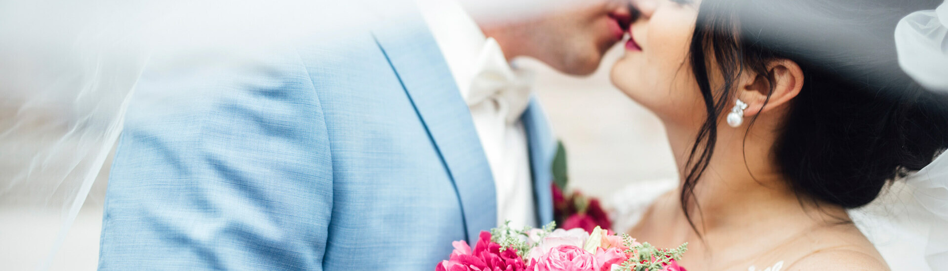 Ein Brautpaar küsst sich, der Bräutigam trägt einen blauen Anzug, die Braut hält einen Blumenstrauß.