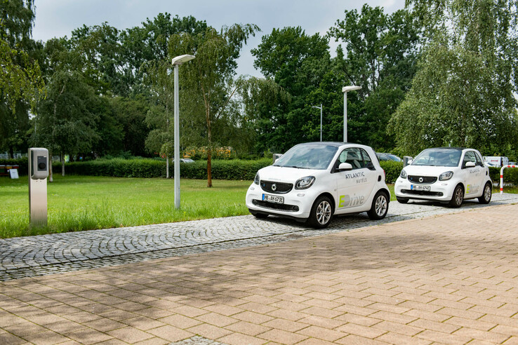 Two white electric cars with the hotel logo at a charging station, surrounded by green parkland.