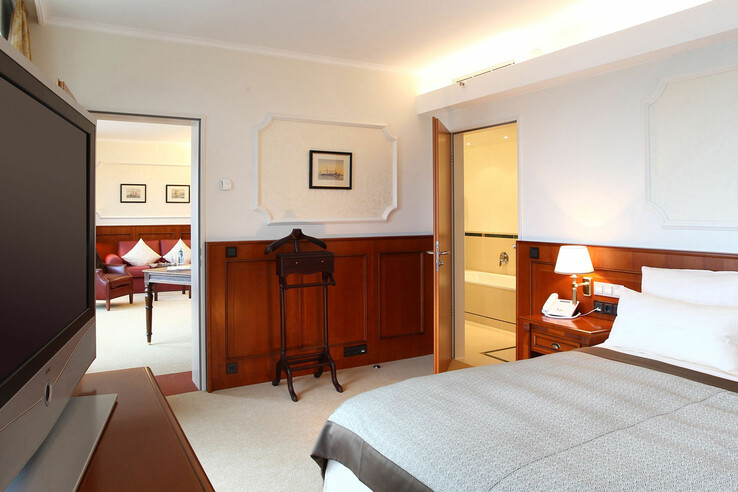 Helles Hotelzimmer mit Doppelbett, TV, Sitzbereich und offenem Badezimmer. Elegante Holzakzente und warme Beleuchtung.