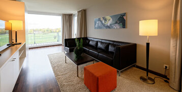 Modern hotel room with leather sofa, large window, view of the racecourse, floor lamp and orange stool on carpet.