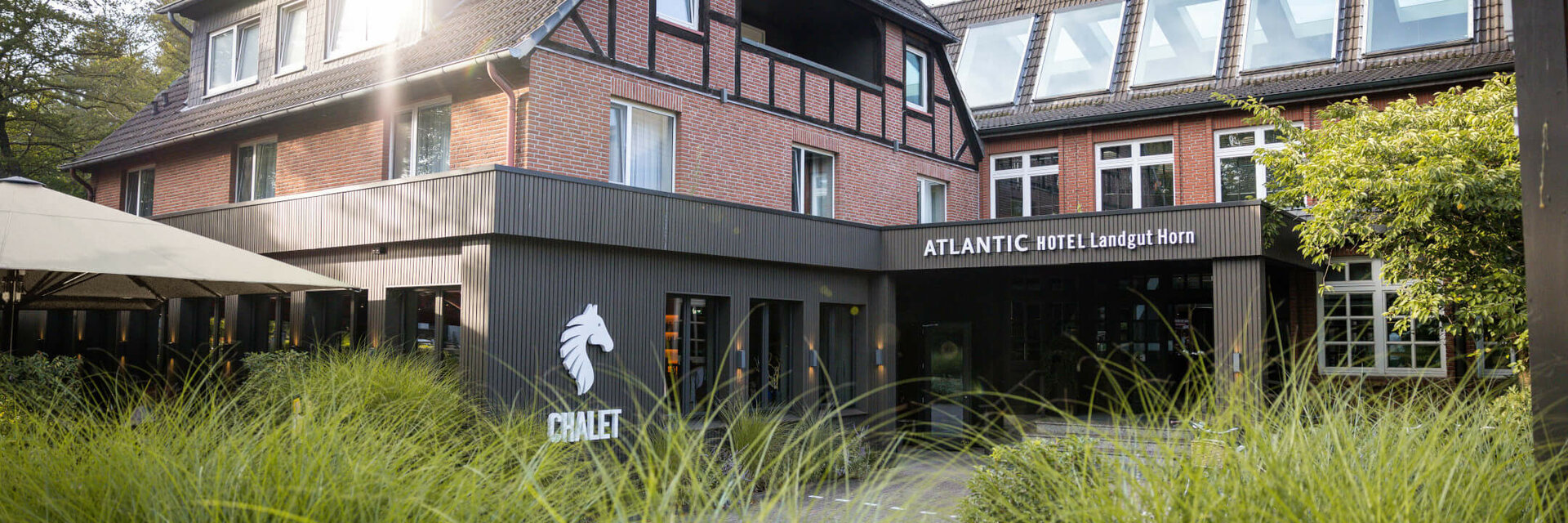 Fachwerkhaus des ATLANTIC Hotel Landgut Horn Bremen mit grünem Garten im Vordergrund und Sonnenschein auf dem Dach.