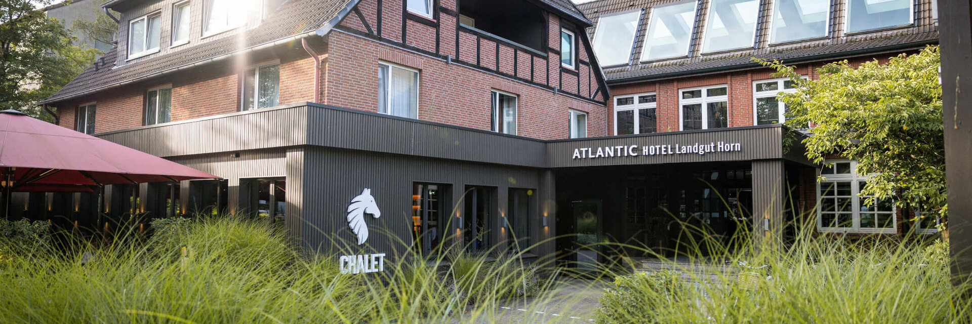 Fachwerkhaus des ATLANTIC Hotel Landgut Horn Bremen mit grünem Garten im Vordergrund und Sonnenschein auf dem Dach.