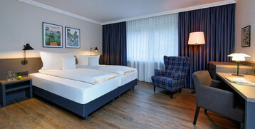 Helles Hotelzimmer mit Doppelbett, Schreibtisch, Sessel, Holzboden und großen Fenstern mit Vorhängen im ATLANTIC Hotel.