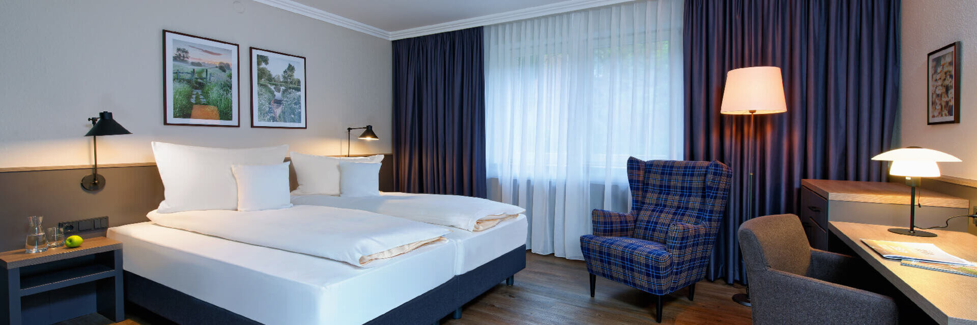 Helles Hotelzimmer mit Doppelbett, Schreibtisch, Sessel, Holzboden und großen Fenstern mit Vorhängen im ATLANTIC Hotel.