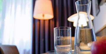 Wasserglas und Karaffe auf Tisch, daneben Lampe und Vorhänge im ATLANTIC Hotel Landgut Horn Bremen.
