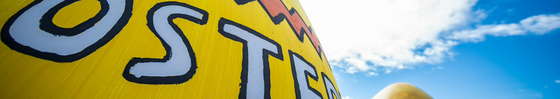 Ostermünde in Travemünde Bunt bemalte Ostereier mit Schriftzug vor blauem Himmel, symbolisieren ein festliches Ereignis.