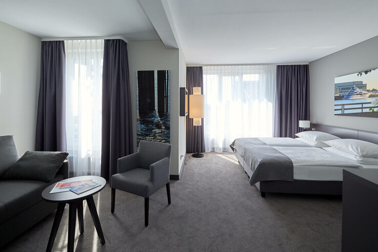 Modernes Hotelzimmer mit Doppelbett, Sofa, Sessel, Tisch und großen Fenstern mit Vorhängen im ATLANTIC Hotel Vegesack.