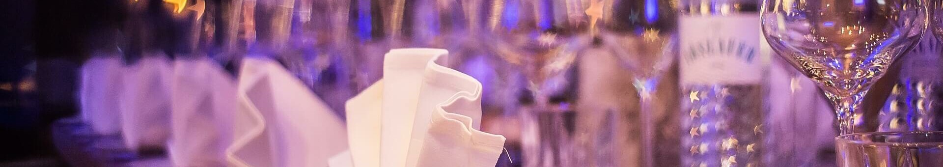 Eleganter, festlich gedeckter Tisch mit gefalteten Servietten, Gläsern und Kerzen in stimmungsvoller Beleuchtung.