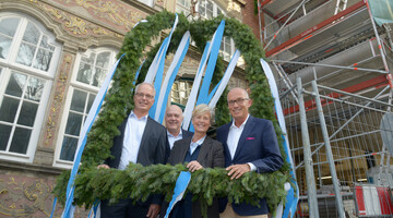 Vier Personen stehen unter einem dekorativen Kranz mit blauen Bändern vor einem historischen Gebäude.