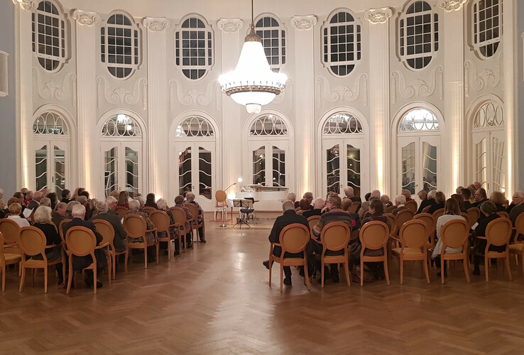 Elegant ballroom in the ATLANTIC Grand Hotel Travemünde with guests, chandelier and high windows.