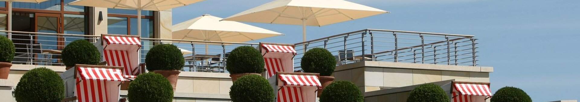 Terrasse mit weißen Sonnenschirmen, roten Strandkörben und grünen Topfpflanzen vor blauem Himmel.