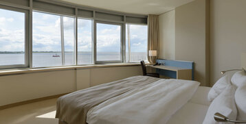 Bright hotel room with large bed, desk and panoramic window with a view of the Weser in Bremerhaven.