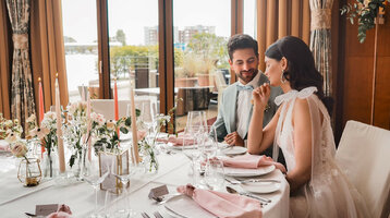 Ein elegantes Paar sitzt an einem festlich gedeckten Tisch mit Blumen und Kerzen im ATLANTIC Hotel Wilhelmshaven.