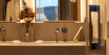 Modernes Badezimmer mit beleuchtetem Spiegel, Waschbecken, Pflegeprodukten und Blumenarrangement im ATLANTIC Hotel Heidelberg.