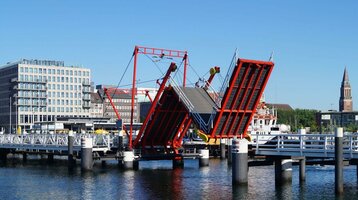 Rote Klappbrücke über Wasser, im Hintergrund moderne Gebäude und Kirchturm bei klarem Himmel.