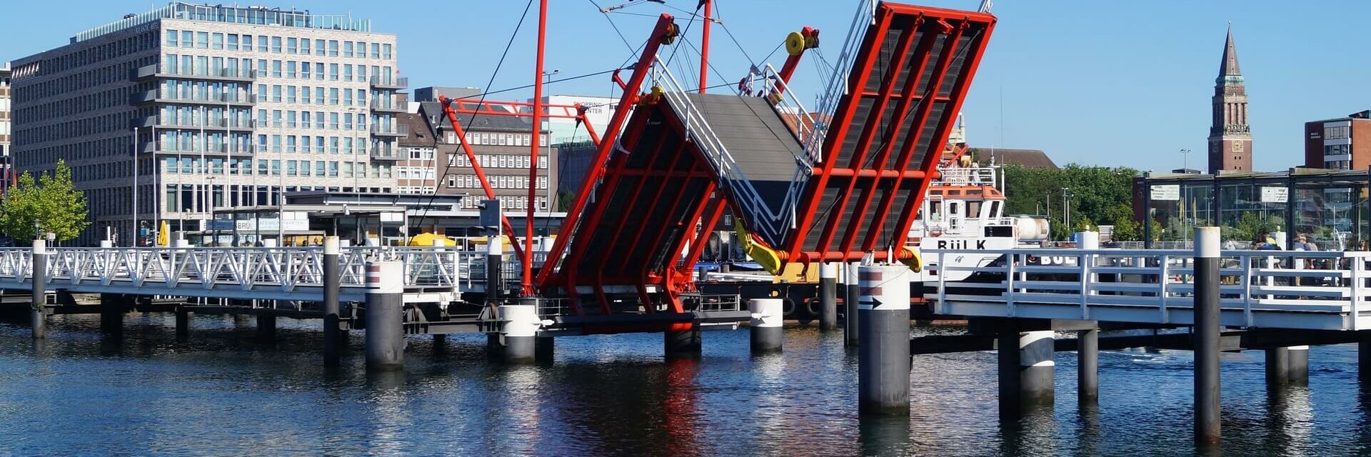 Rote Klappbrücke über Wasser, im Hintergrund moderne Gebäude und Kirchturm bei klarem Himmel.
