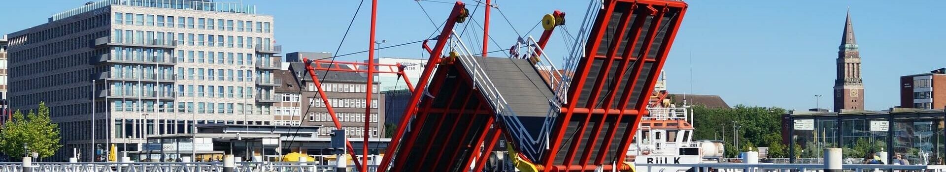 Rote Klappbrücke über Wasser, im Hintergrund moderne Gebäude und Kirchturm bei klarem Himmel.