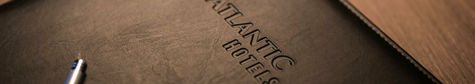 Black leather folder with "ATLANTIC HOTELS" embossing on a wooden table, with a silver ballpoint pen next to it.