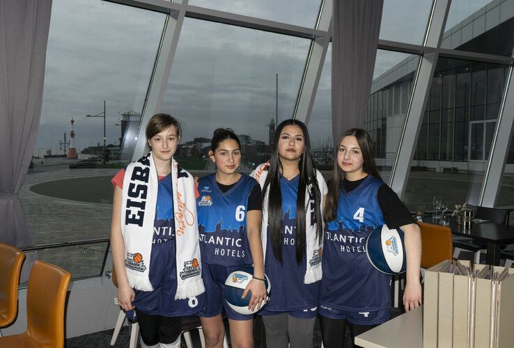 Four basketball players in ATLANTIC Hotels jerseys pose in front of a window with a view of the harbor.